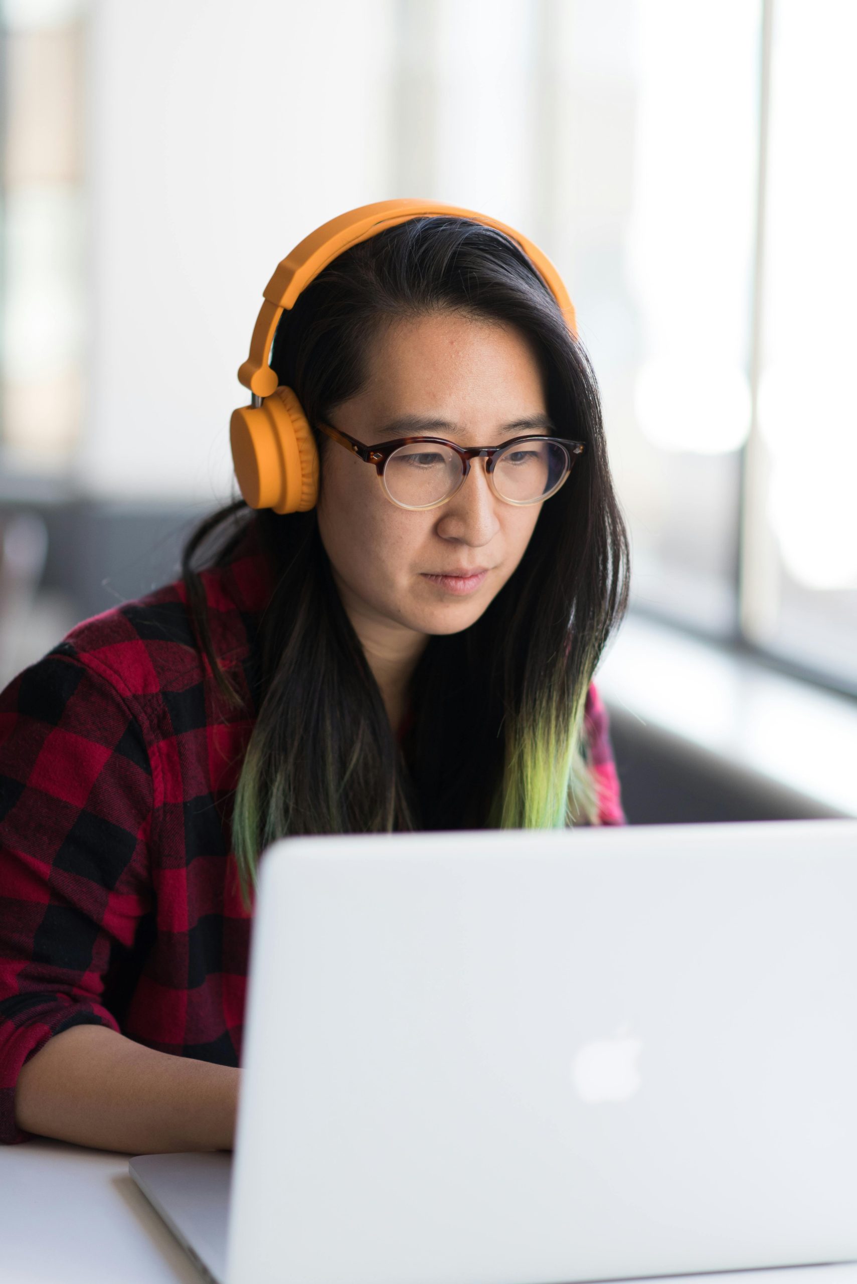 Focused Asian woman using laptop with headphones in a bright room, epitomizing remote work.